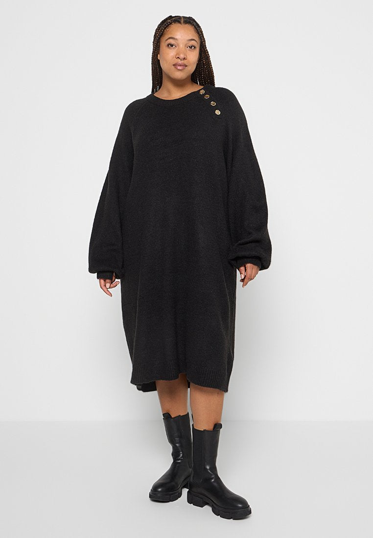 Schwarzes, oversize Strickkleid mit langen Ärmeln, Rundhalsausschnitt und drei dekorativen Knöpfen an der Schulter. Getragen mit kniehohen schwarzen Stiefeln.