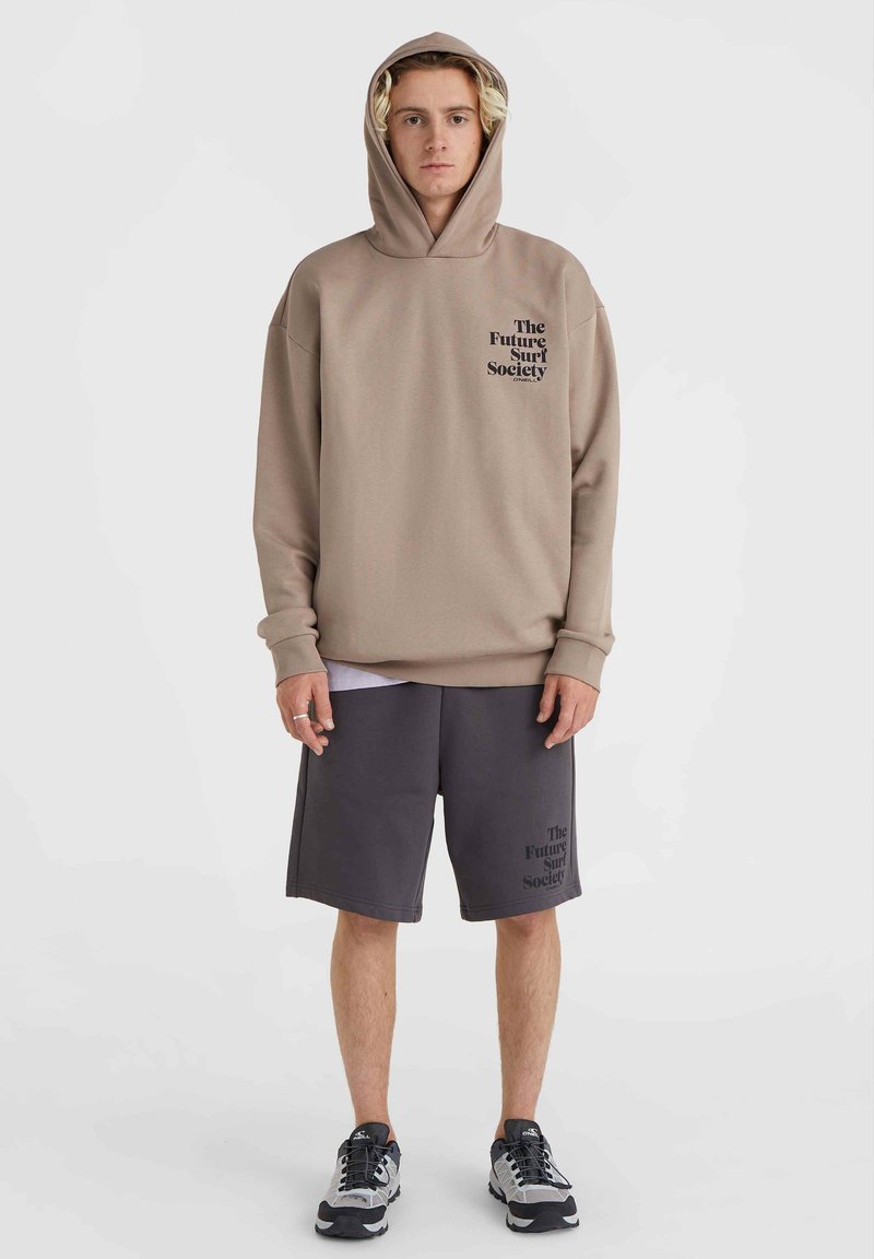 Beige Kapuzenpullover mit einem Frontlogo, kombiniert mit dunkelgrauen Shorts, die dasselbe Logo zeigen. Das Modell trägt graue Sneakers.