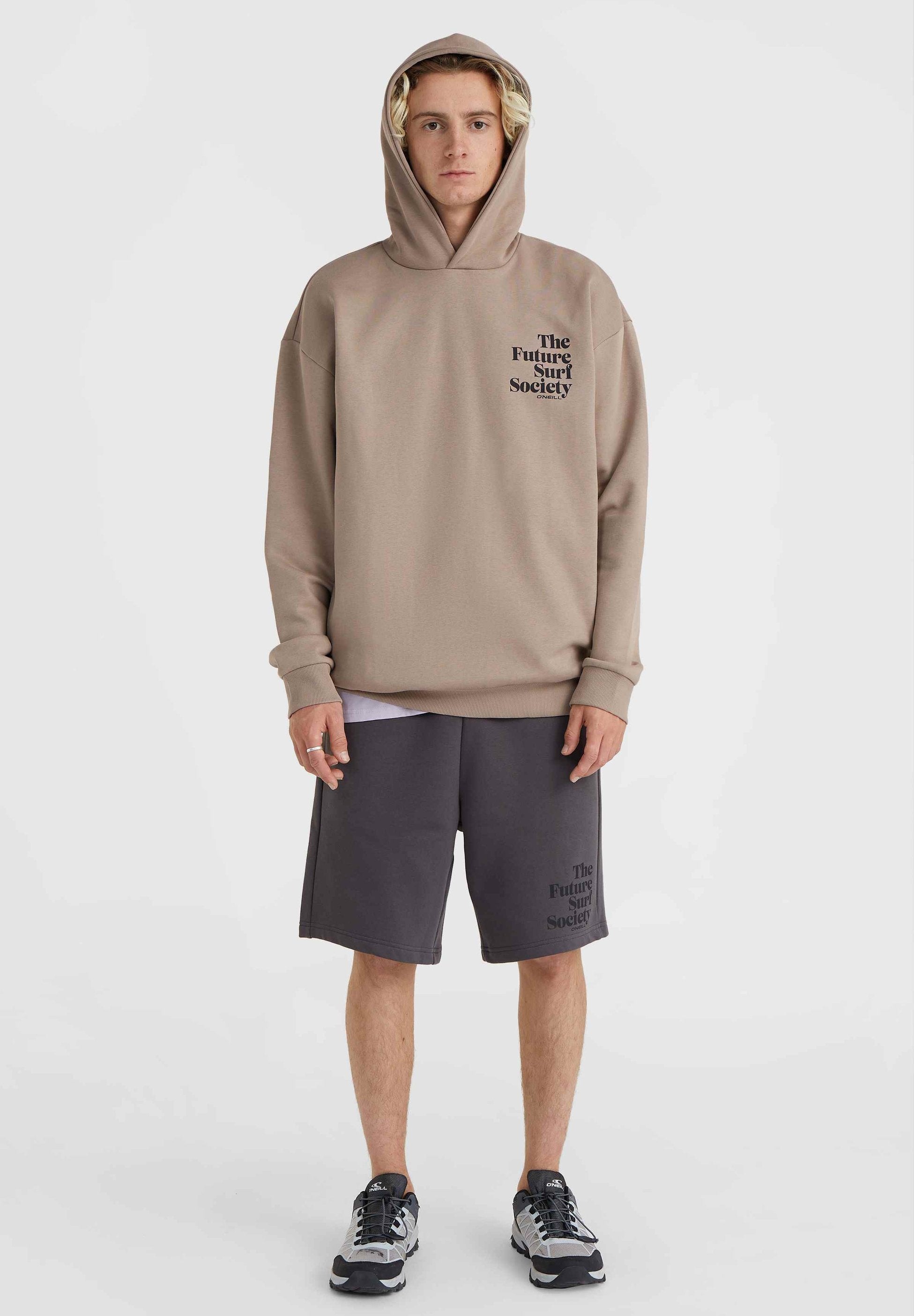 Beige Kapuzenpullover mit einem Frontlogo, kombiniert mit dunkelgrauen Shorts, die dasselbe Logo zeigen. Das Modell trägt graue Sneakers.
