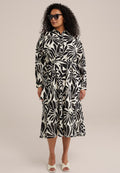 Schwarz-weißes Kleid mit Blättermuster, kragenloser Hals, langen Ärmeln, Taillengürtel und gestuftem Saum. Getragen mit hellen, hohen Sandalen.