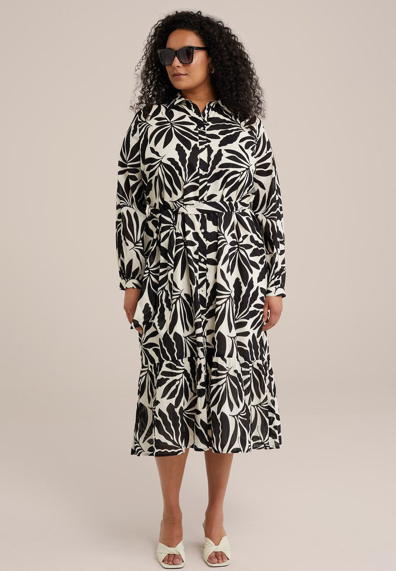 Schwarz-weißes Kleid mit Blättermuster, kragenloser Hals, langen Ärmeln, Taillengürtel und gestuftem Saum. Getragen mit hellen, hohen Sandalen.