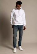 Mann, der einen weißen Kapuzenpullover, blaue, ausgewaschene Jeans und weiße Sneakers trägt, steht vor einem schlichten beigefarbenen Hintergrund und schaut nach unten.