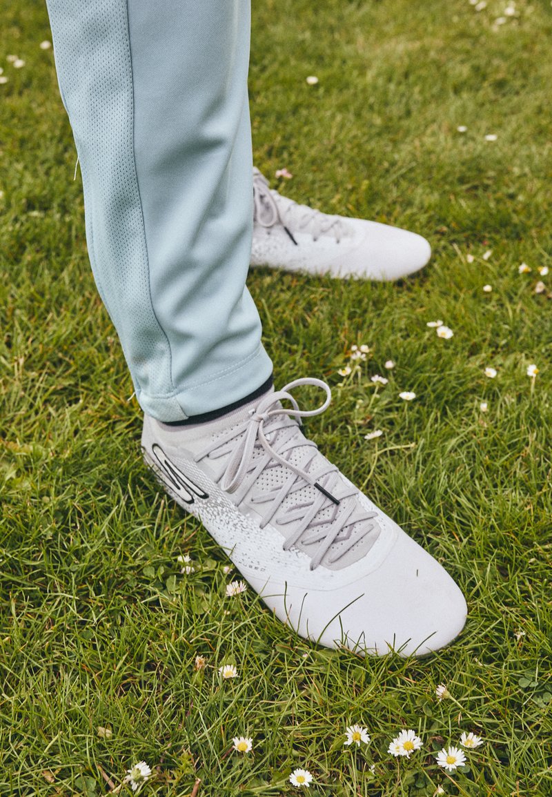 Hellgraue Sportschuhe mit strukturierten Flächen und Schnürsenkeln, kombiniert mit hellblauen Sporthosen, die auf einem mit kleinen Blumen bewachsenen Grasboden stehen.