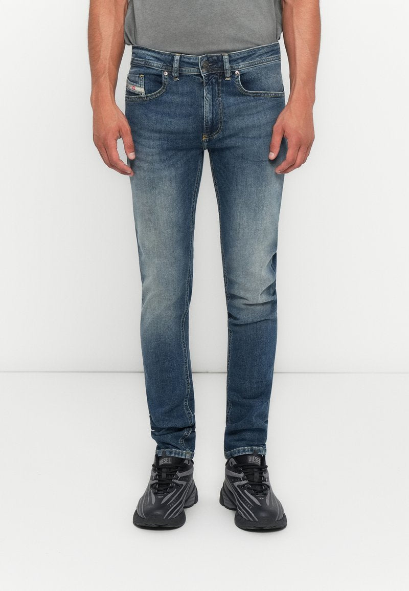 Mann in verwaschenen blauen Slim-Fit-Jeans und schwarzen Diesel-Sneakern, der vor einem weißen Hintergrund steht.
