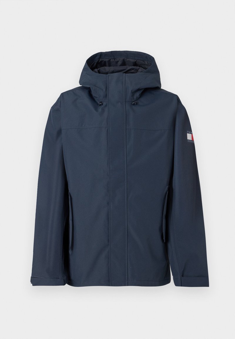 Marineblaue wasserdichte Jacke mit Kapuze, Front-Reißverschluss, Seitentaschen und Logodetail am Ärmel. Glatte Textur und modernes Design.