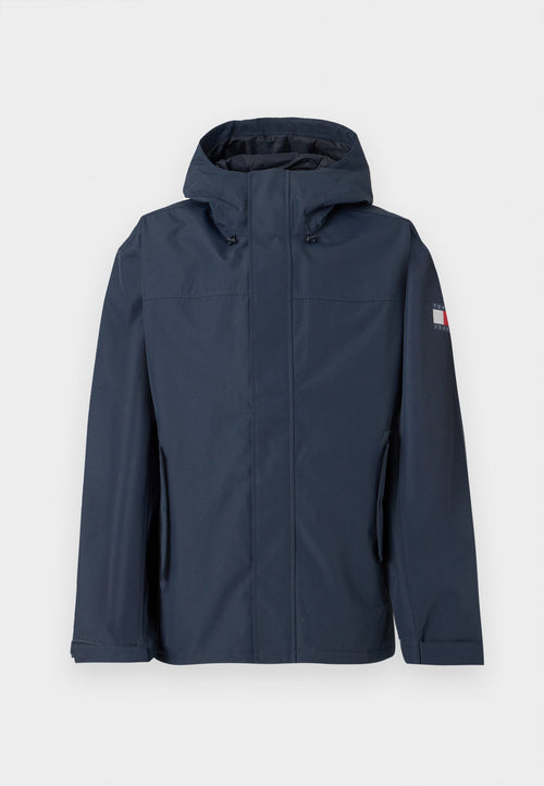Marineblaue wasserdichte Jacke mit Kapuze, Front-Reißverschluss, Seitentaschen und Logodetail am Ärmel. Glatte Textur und modernes Design.