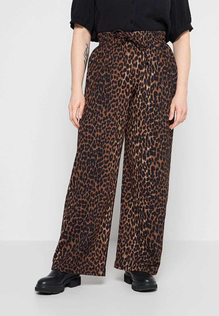 Person trägt braune und schwarze Leopardenmuster-Hose mit weitem Bein und Paperbag-Taille sowie schwarze klobige Stiefel, steht vor einem schlichten Hintergrund.