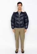 Ein Mann steht mit dem Gesicht nach vorne und trägt eine marineblaue Steppjacke mit weißem Schafswollkragen, beigefarbene Cargo-Hosen und beigefarbene Wanderschuhe vor einem weißen Hintergrund.