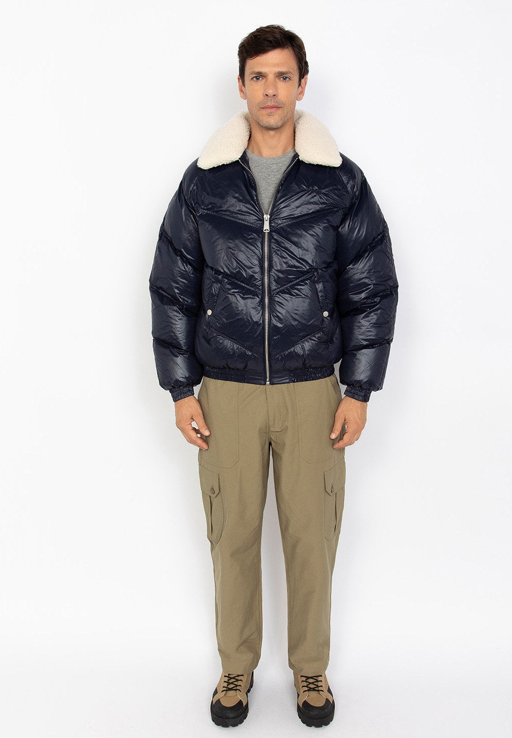 Ein Mann steht mit dem Gesicht nach vorne und trägt eine marineblaue Steppjacke mit weißem Schafswollkragen, beigefarbene Cargo-Hosen und beigefarbene Wanderschuhe vor einem weißen Hintergrund.