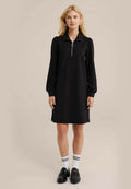 Schwarzes Kleid mit Kragen und Reißverschluss, mit langen Ärmeln und lockerem Schnitt. Getragen mit schwarzen Slippern und weißen Socken mit schwarzen Streifen.