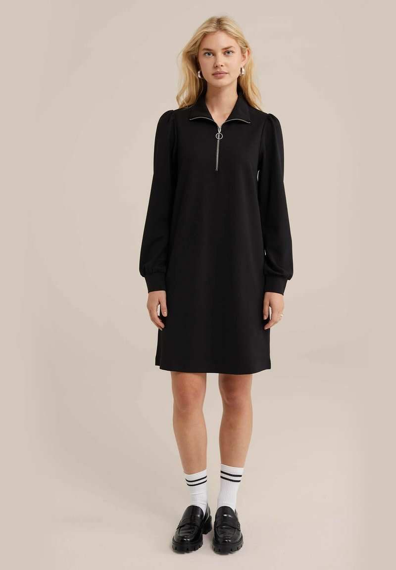Schwarzes Kleid mit Kragen und Reißverschluss, mit langen Ärmeln und lockerem Schnitt. Getragen mit schwarzen Slippern und weißen Socken mit schwarzen Streifen.