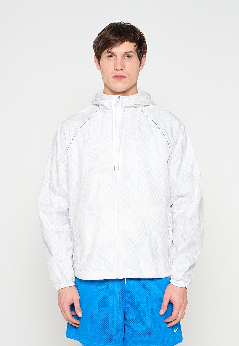 Junger Mann mit weißem, gemustertem Kapuzen-Windbreaker und leuchtend blauen, atmungsaktiven Sportshorts vor einfarbigem Hintergrund.
