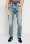 Mann in hellblauen, ausgebleichten Jeans, schwarzen Sneakers mit weißen Streifen und einem braun gestreiften Hemd, der vor einem weißen Hintergrund steht.