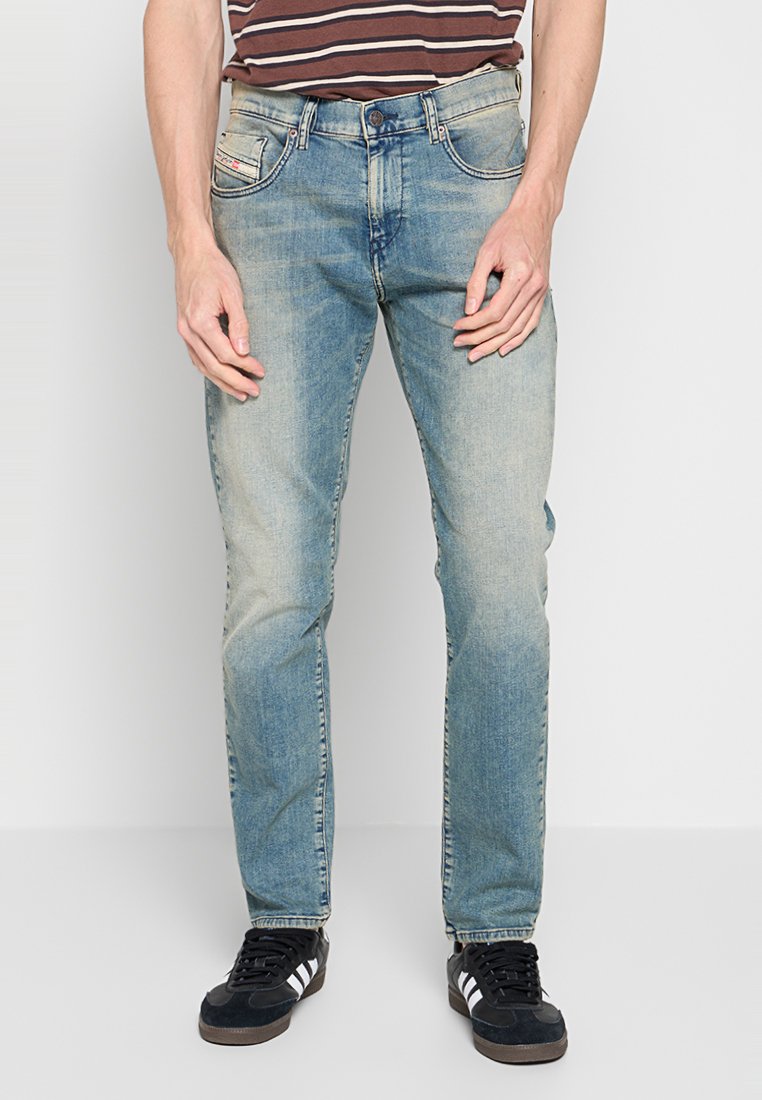 Mann in hellblauen, ausgebleichten Jeans, schwarzen Sneakers mit weißen Streifen und einem braun gestreiften Hemd, der vor einem weißen Hintergrund steht.