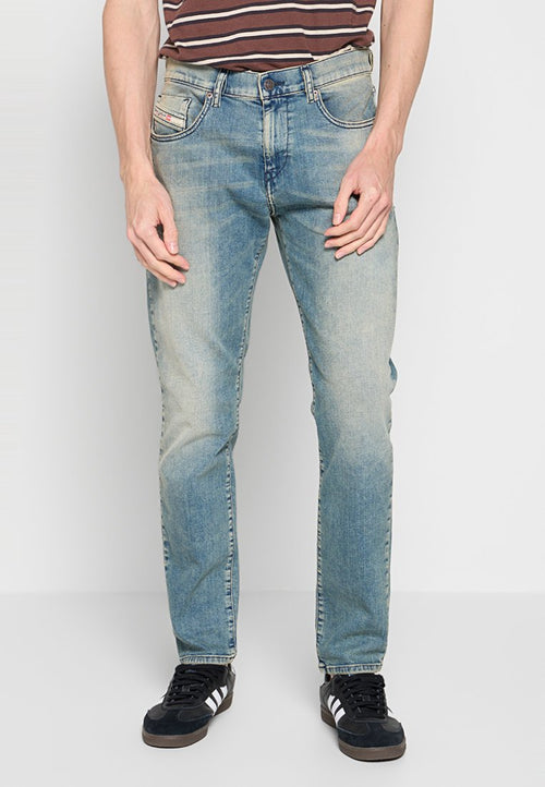 Mann in hellblauen, ausgebleichten Jeans, schwarzen Sneakers mit weißen Streifen und einem braun gestreiften Hemd, der vor einem weißen Hintergrund steht.