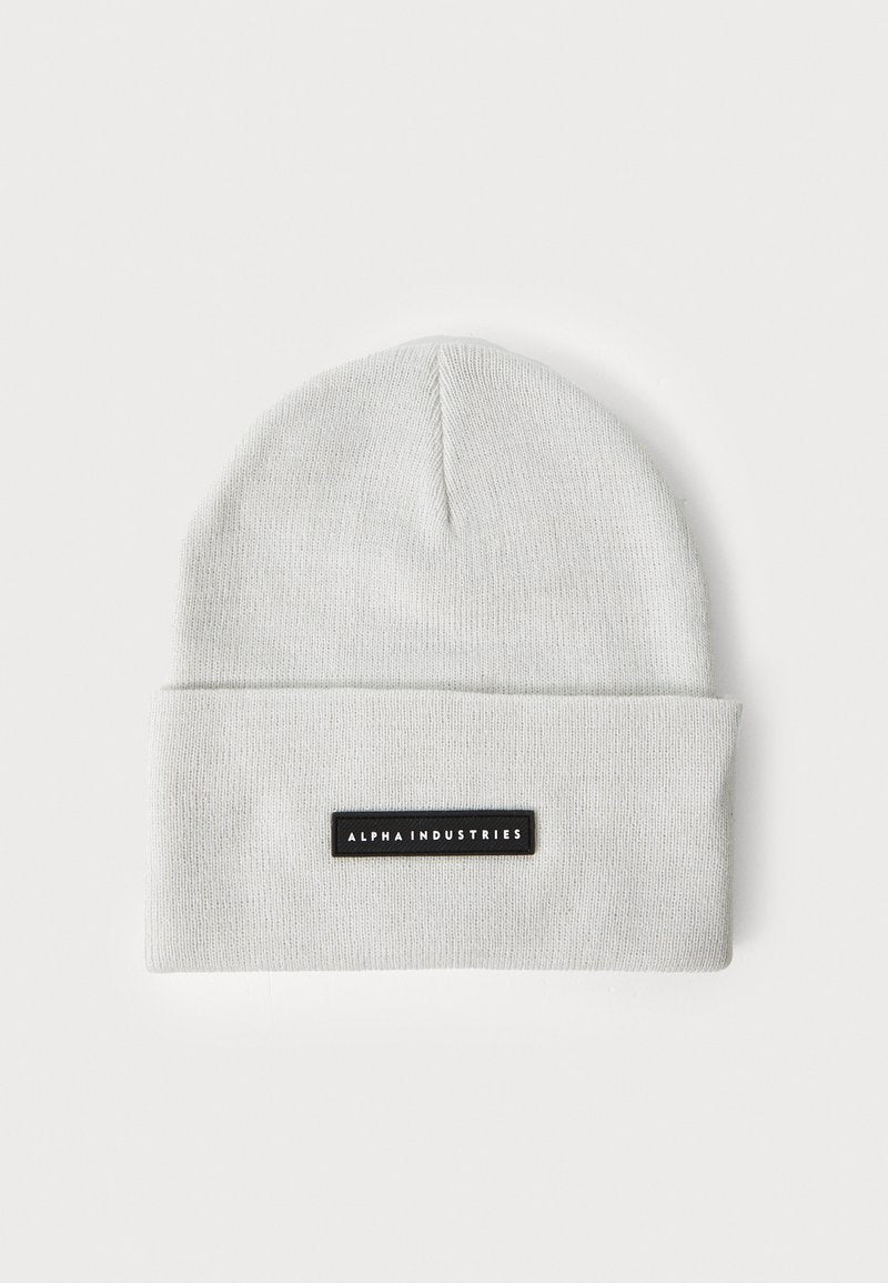 Hellgraue Strickmütze mit umgeschlagenem Bündchen und einem rechteckigen schwarzen Etikett mit weißem Text für "ALPHA INDUSTRIES". Glatte Textur.