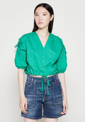 Grüne, kürzer geschnittene Bluse mit V-Ausschnitt, Puffärmeln, Knöpfen vorne und Kordelzug in der Taille, kombiniert mit blauen Jeansshorts. Glatte Textur.