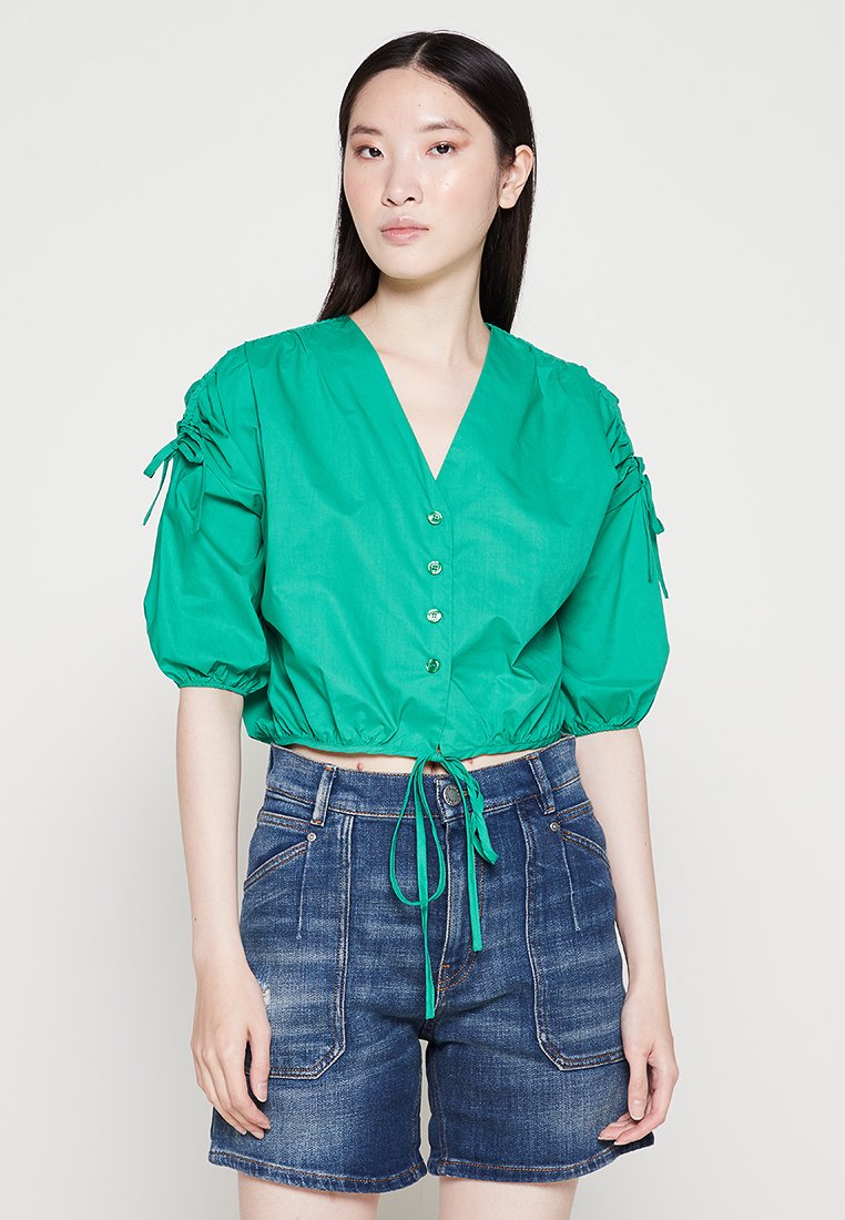 Grüne, kürzer geschnittene Bluse mit V-Ausschnitt, Puffärmeln, Knöpfen vorne und Kordelzug in der Taille, kombiniert mit blauen Jeansshorts. Glatte Textur.
