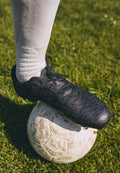 Schwarzer Fußballschuh mit strukturiertem, rissigem Design, geschnürt an einem Fuß über einem schmutzigen weißen Fußball auf Gras. Weiße Crew-Socke sichtbar.
