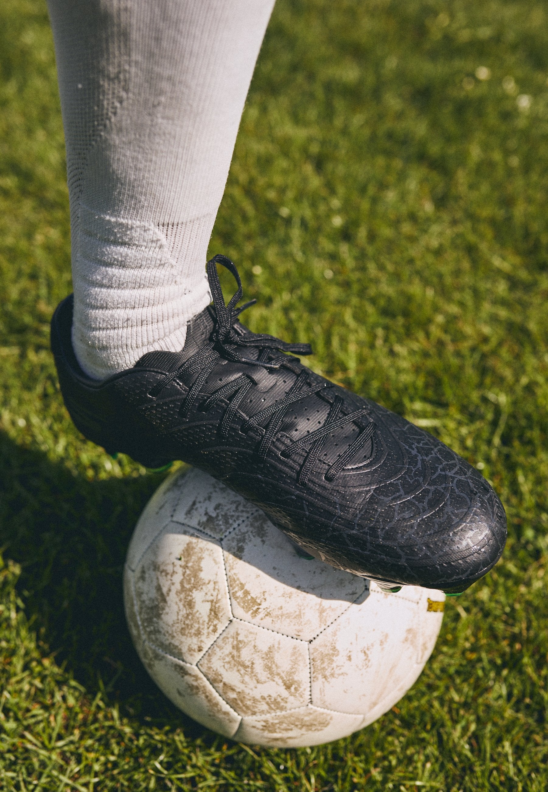 Schwarzer Fußballschuh mit strukturiertem, rissigem Design, geschnürt an einem Fuß über einem schmutzigen weißen Fußball auf Gras. Weiße Crew-Socke sichtbar.