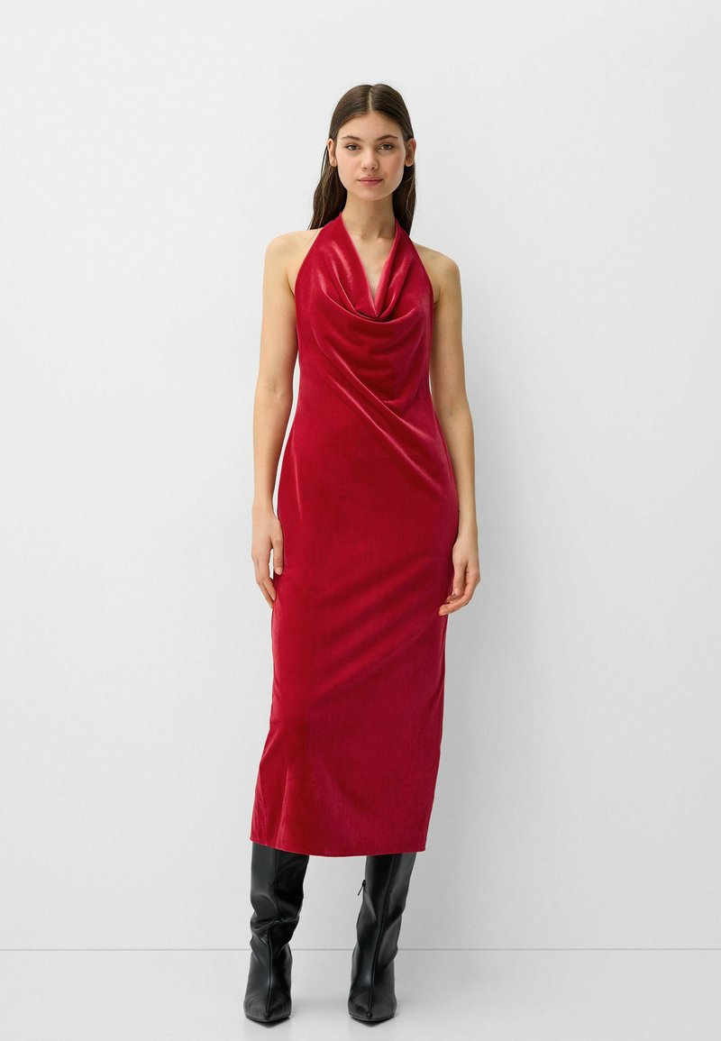 Ein rotes Samt-Halterneck-Kleid mit schmalem Ausschnitt, bodenlangem Design und glatter Textur, kombiniert mit schwarzen kniehohen Stiefeln.