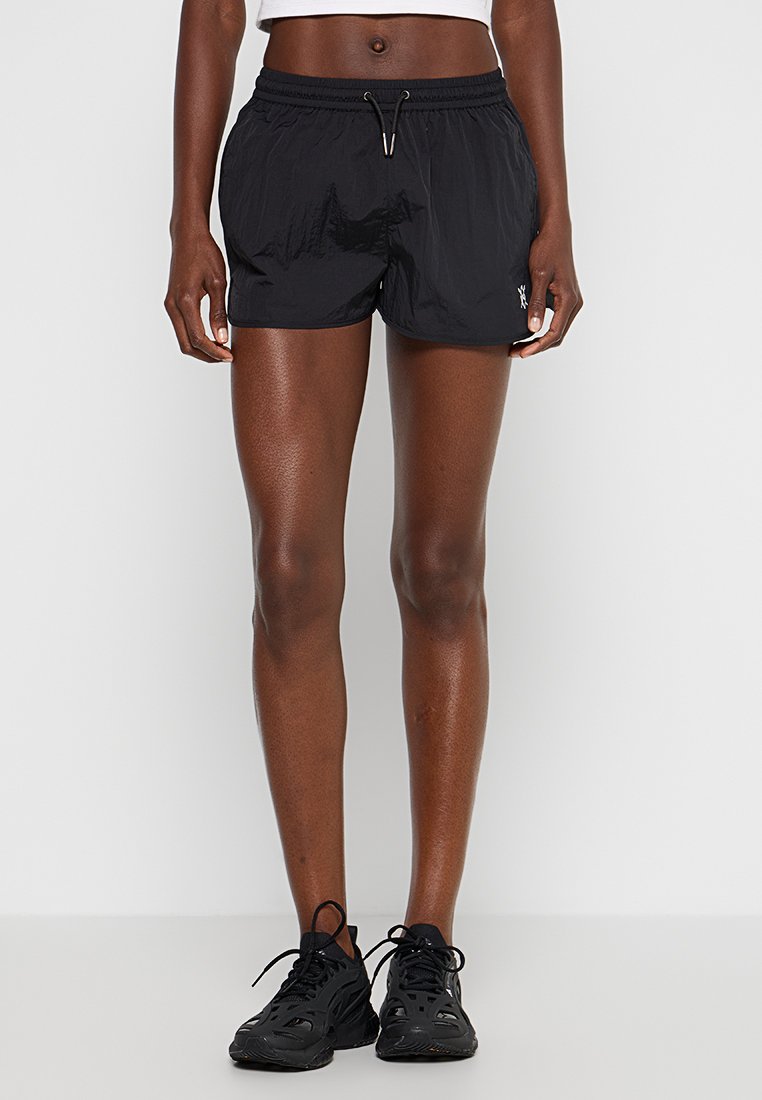 Schwarze Sportshorts mit glatter Textur, elastischem Bund und Kordelzug. Logobezug an der unteren linken Ecke. Getragen mit schwarzen Sneakers.