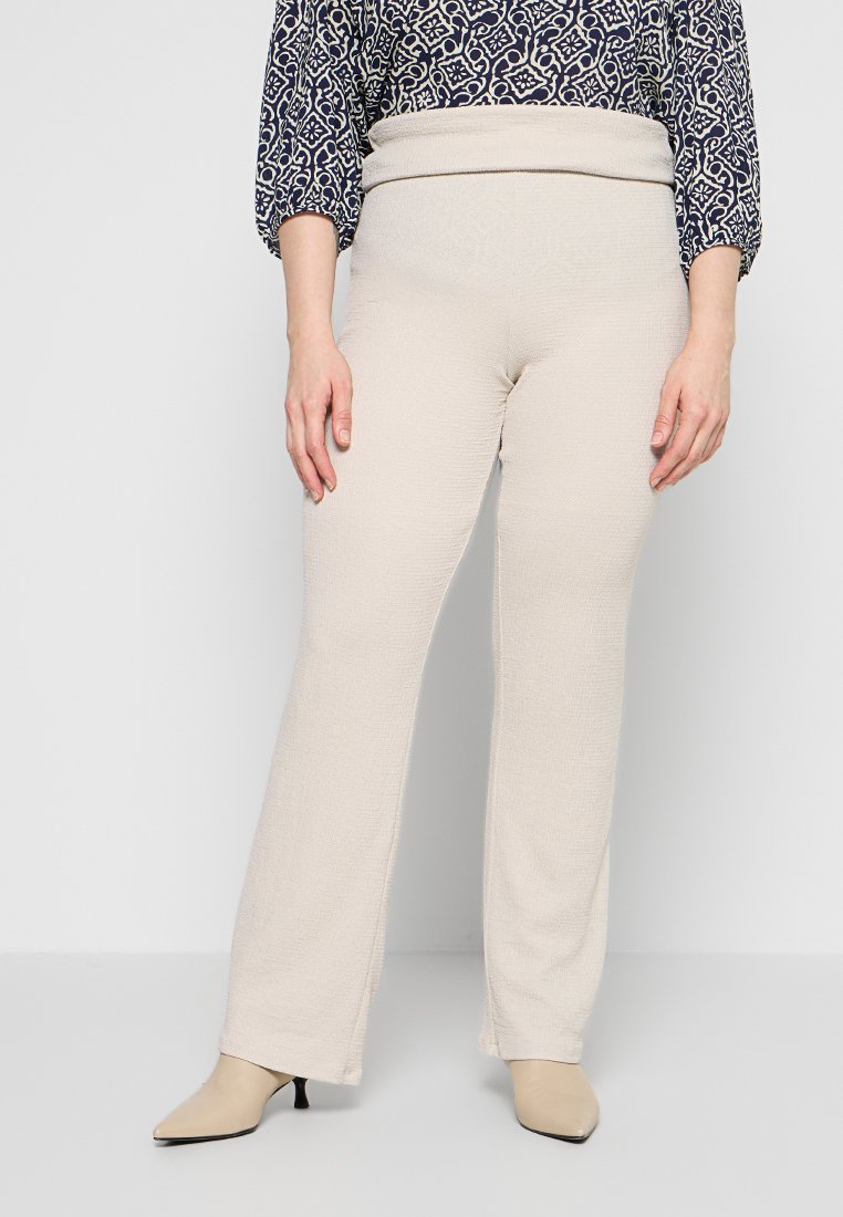 Frau trägt beige Textilhose mit hohem Bund und umgeschlagener Taille, kombiniert mit beigen spitzen Pumps und einer gemusterten Bluse.