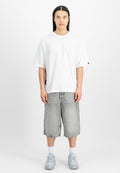 Junge Person mit langen schwarzen Haaren, die ein schlichtes weißes übergroßes T-Shirt, graue knielange Jeansshorts, weiße Socken und silberne Turnschuhe trägt.