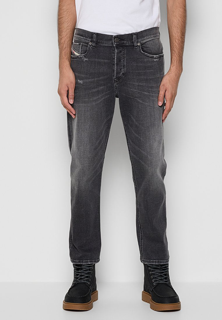 Mann in ausgebleichten schwarzen Jeans mit leichtem Used-Look und schwarzen Schnürstiefeln mit braunen Sohlen, entspannt mit den Händen stehend.