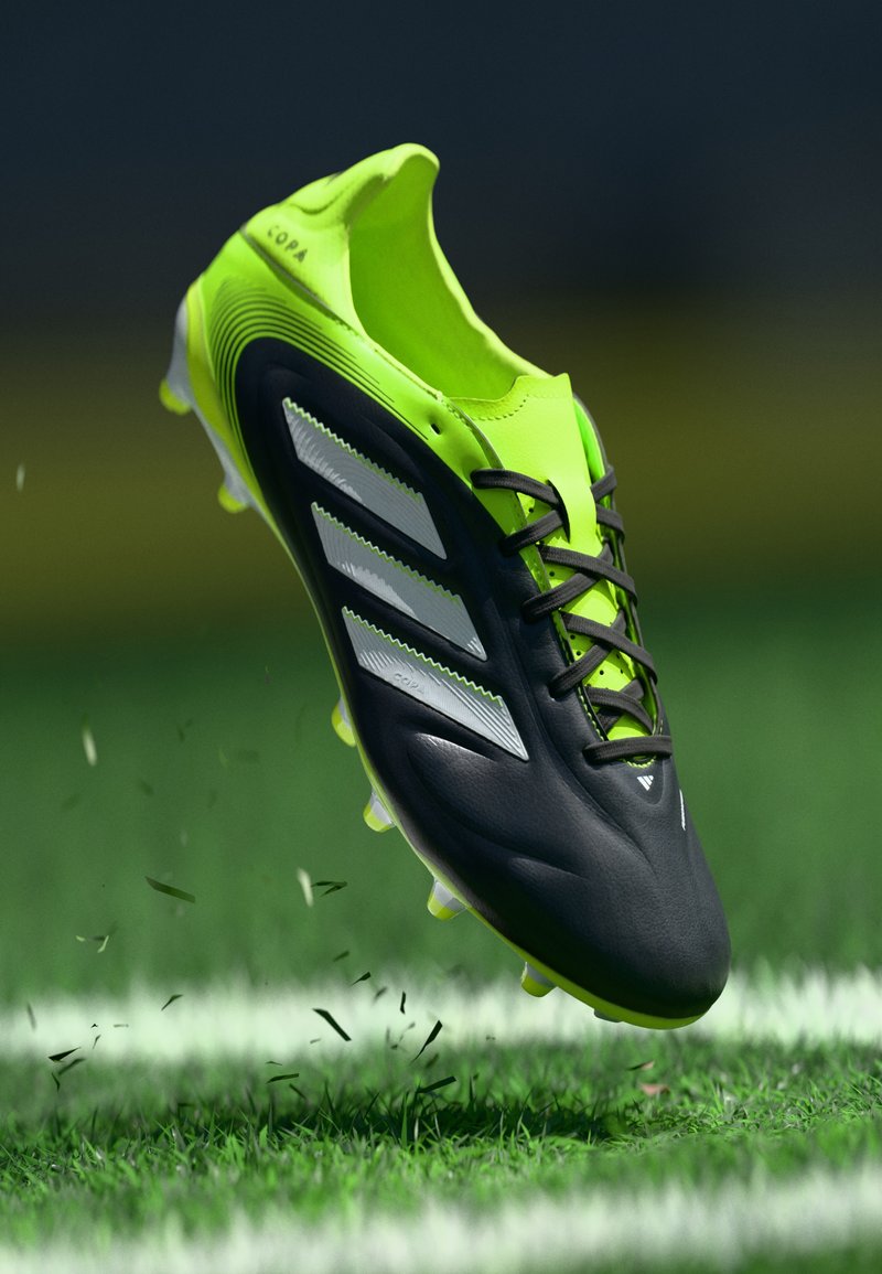Schwarzer und neongrüner Fußballschuh mit drei weißen Streifen, strukturiertem Obermaterial, Schnürdesign und gelben Stollen, schwebend über Gras.