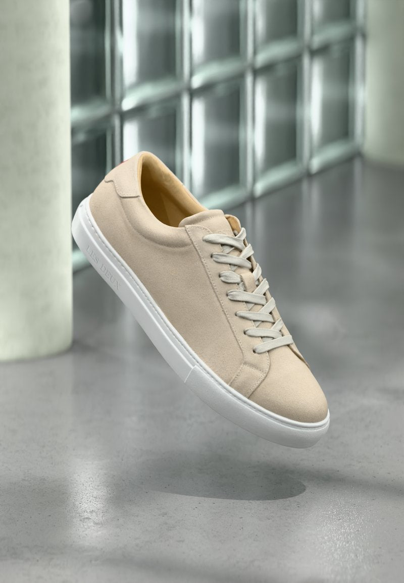 Beige Wildleder-Sneaker mit weißer Gummisohle und Schnürsenkeln. Sie verfügen über eine glatte Textur und ein minimalistisches Design, das vor einem neutralen Hintergrund schwebt.