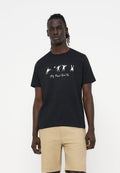 Mann trägt schwarzes T-Shirt mit weißen abstrakten Figuren und Text, kombiniert mit beigen Shorts vor einem schlichten weißen Hintergrund.