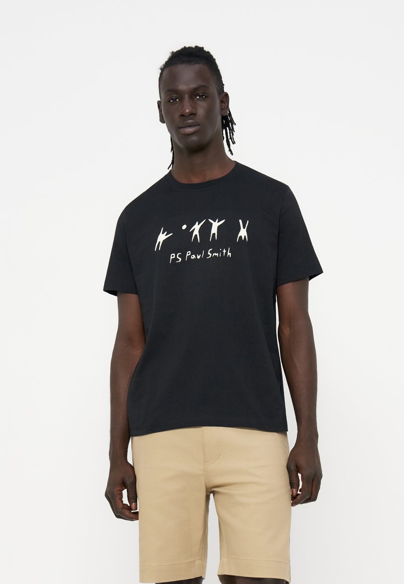 Mann trägt schwarzes T-Shirt mit weißen abstrakten Figuren und Text, kombiniert mit beigen Shorts vor einem schlichten weißen Hintergrund.