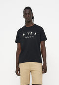 Mann trägt schwarzes T-Shirt mit weißen abstrakten Figuren und Text, kombiniert mit beigen Shorts vor einem schlichten weißen Hintergrund.