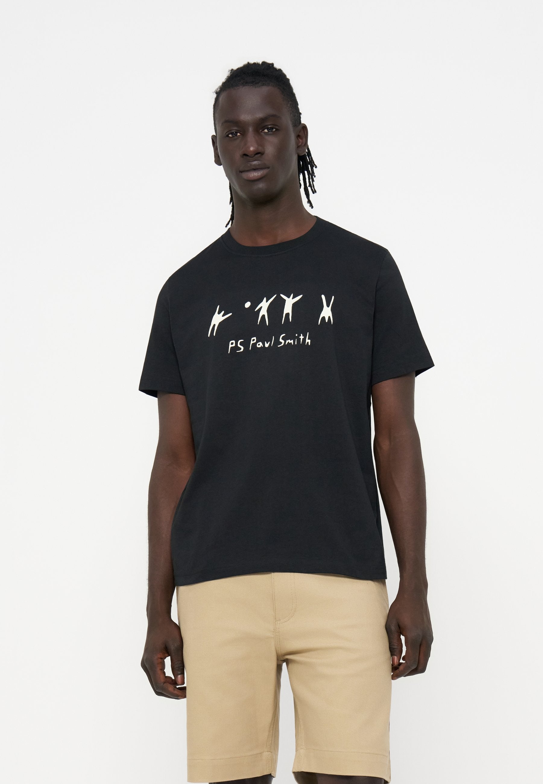 Mann trägt schwarzes T-Shirt mit weißen abstrakten Figuren und Text, kombiniert mit beigen Shorts vor einem schlichten weißen Hintergrund.