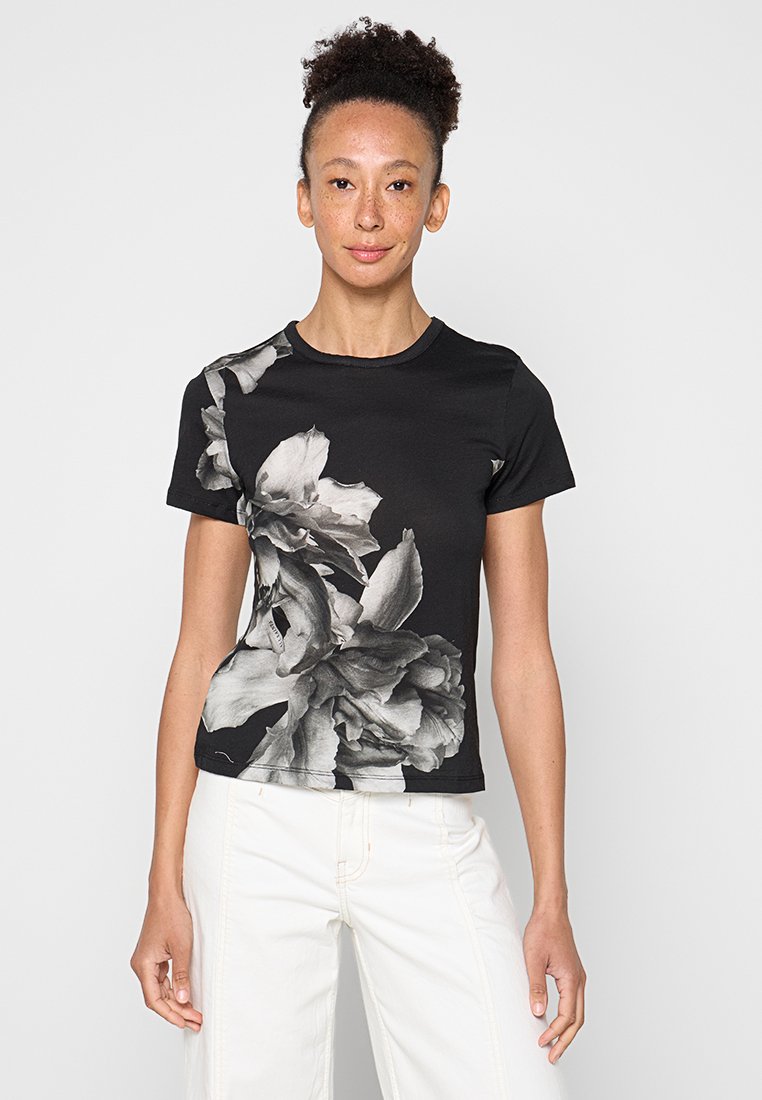 Frau trägt ein schwarzes T-Shirt mit kurzen Ärmeln und großem Graustufen-Blumenmuster und weiße Hose, steht vor einfarbigem Hintergrund.