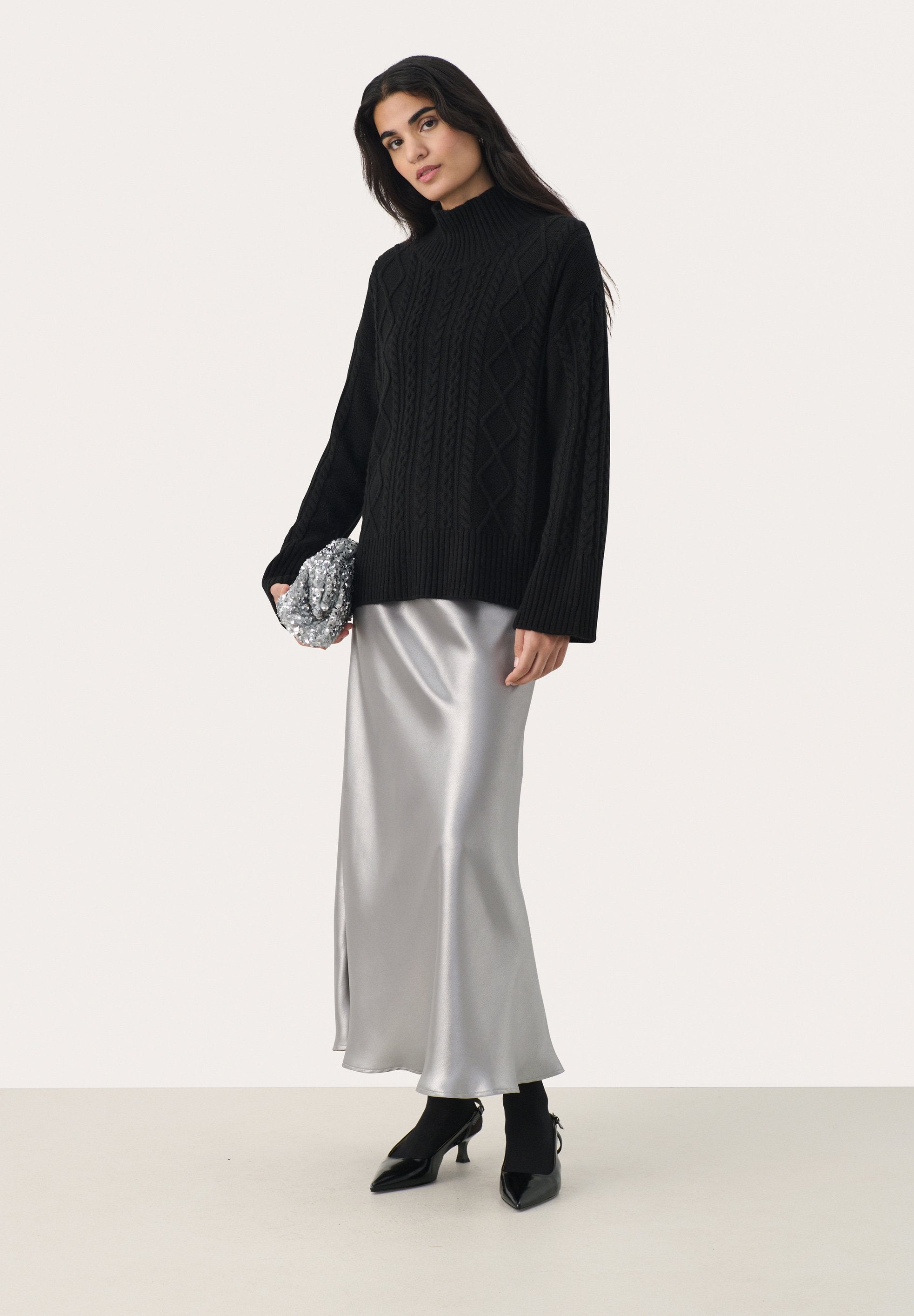 Schwarzer Strickpullover mit hohem Kragen, kombiniert mit einem silbernen Satinrock mit seitlichem Schlitz. Das Model hält eine strukturierte silberne Clutch.