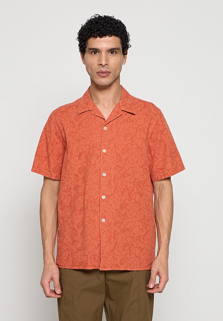 Mann mit lockigem Haar, der ein orangefarbenes kurzärmeliges Hemd mit subtilem Blumenmuster und braune Hosen trägt, steht vor einem einfarbigen Hintergrund.