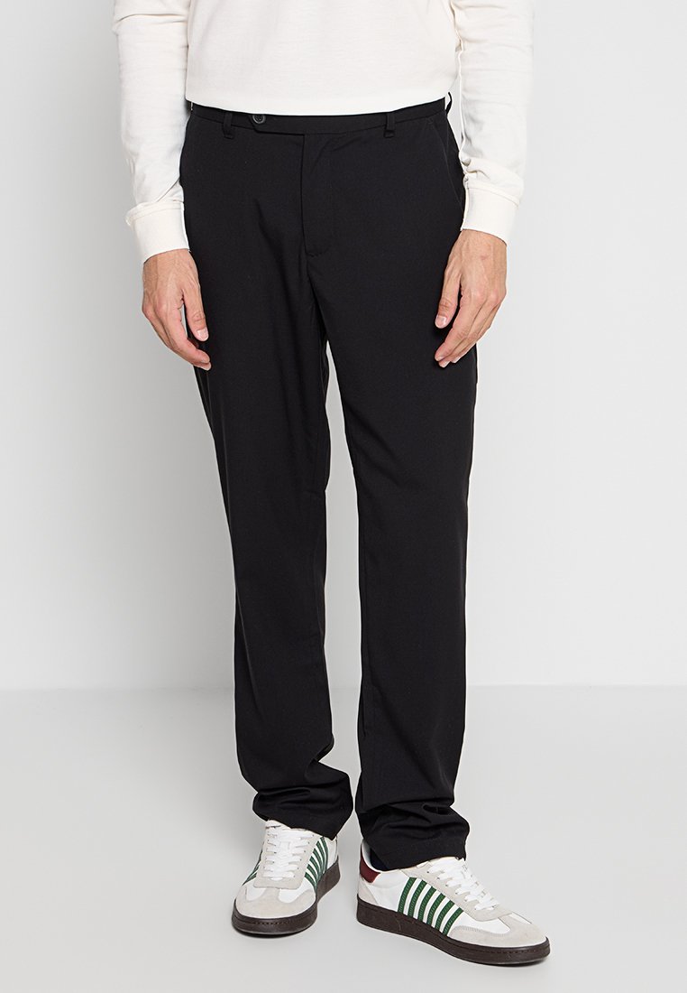Schwarze maßgeschneiderte Hose mit glatter Textur, mit Knopfverschluss und Gürtelschlaufen, kombiniert mit grau-weißen Sneakers mit grünen Streifen.