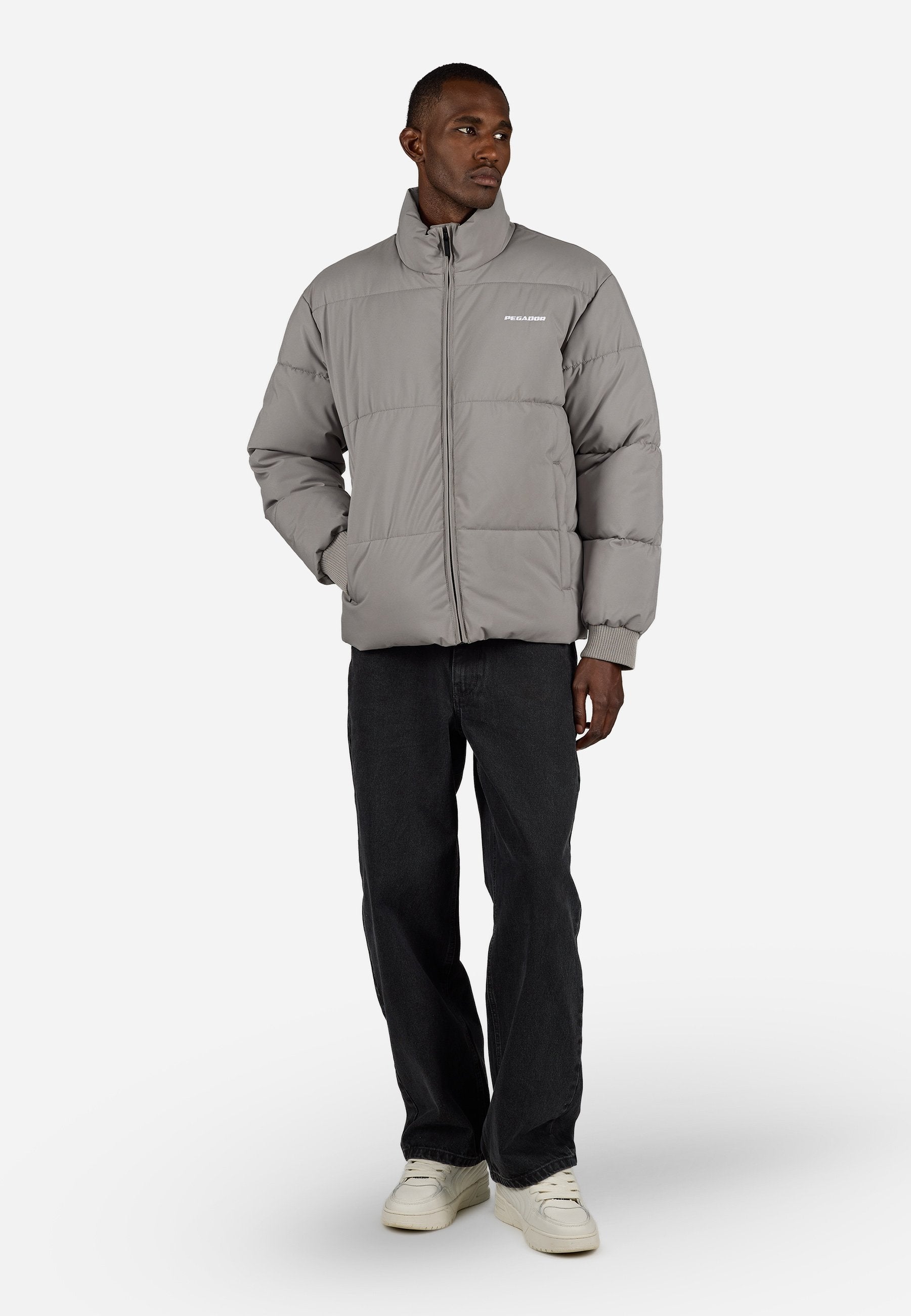 Graue Steppjacke mit hohem Kragen, Reißverschluss und gerippten Bündchen. Logo auf der Brust gedruckt. Kombiniert mit schwarzen Jeans und weißen Sneakers.