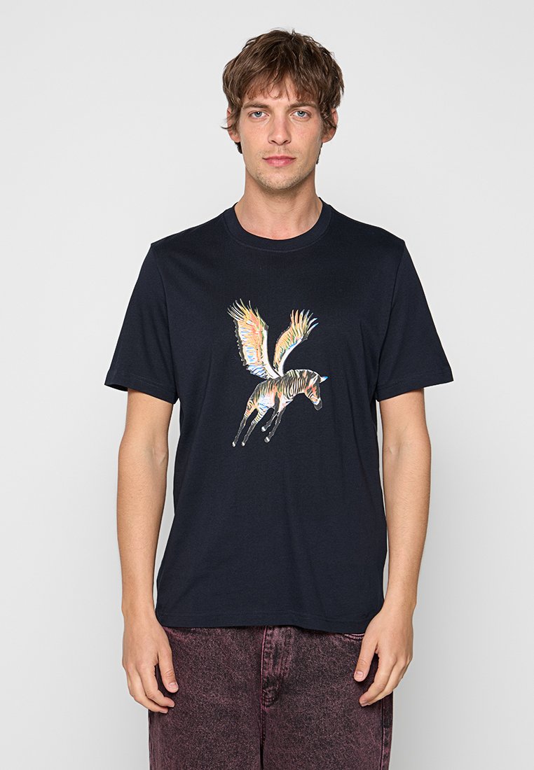 Mann trägt ein schwarzes T-Shirt mit einem bunten, geflügelten Zebra-Motiv, kombiniert mit dunkelroten, gewaschenen Hosen, vor einem schlichten hellen Hintergrund.