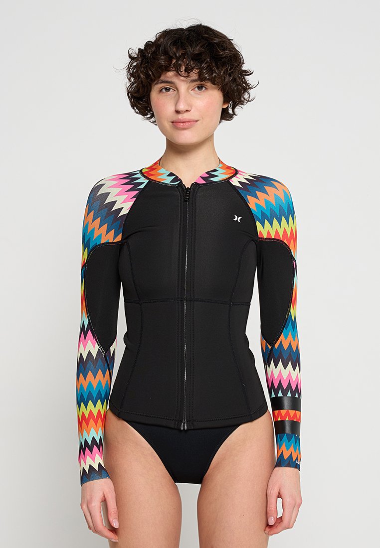 Frau in einer schwarzen Neoprenjacke mit bunten Zickzack-Mustern an den Ärmeln und einem Reißverschluss vorne, steht vor einem schlichten Hintergrund.