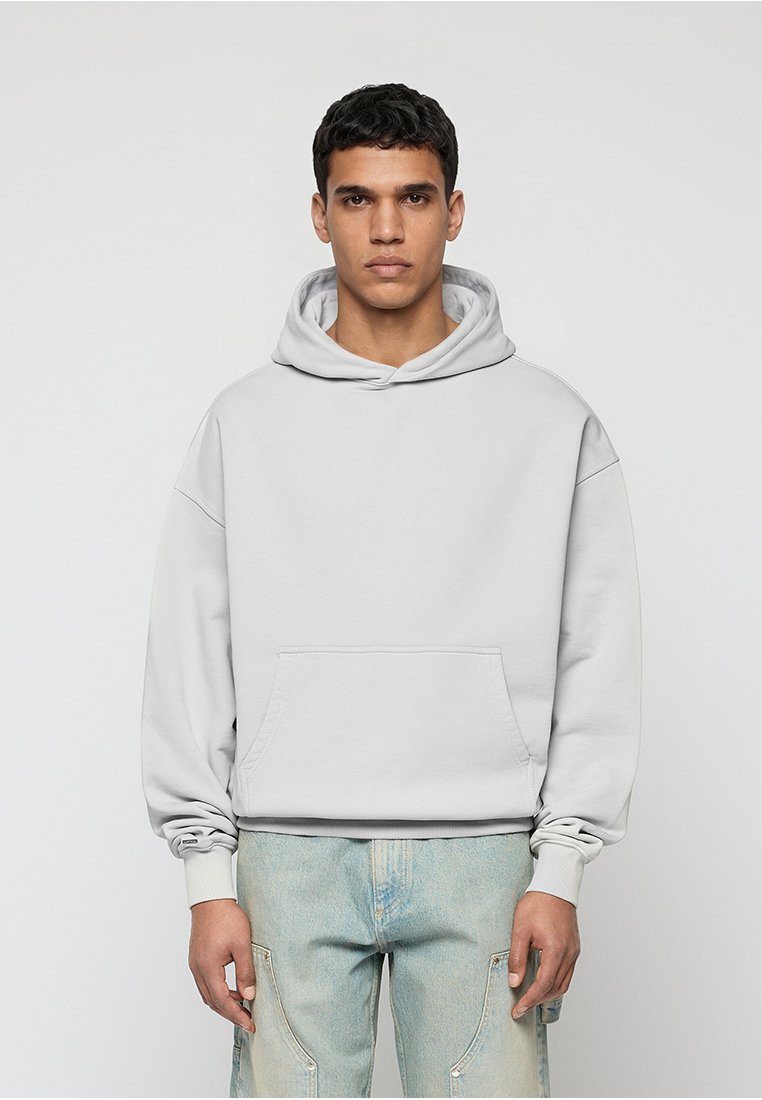 Junger Mann, der frontal steht, trägt einen schlichten hellgrauen Kapuzenpullover und hellblaue Jeans vor einem einfarbigen Hintergrund.
