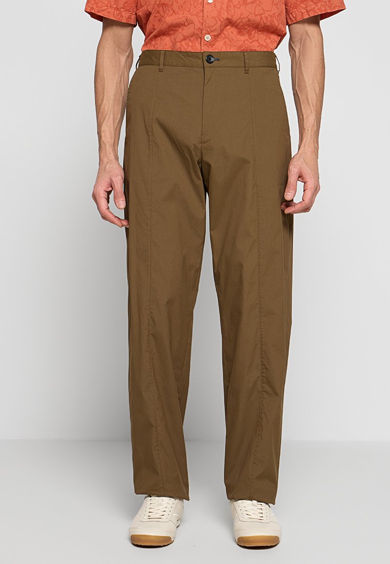 Mann trägt braune gerade geschnittene Hose, weiße Turnschuhe und ein orangefarbenes kurzärmeliges Hemd mit dezentem Muster.