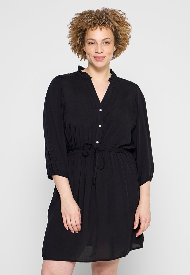 Frau mit lockigem Haar, die ein schwarzes, knöpfgemachtes Kleid mit gebundener Taille trägt und vor einem einfarbigen hellen Hintergrund steht.