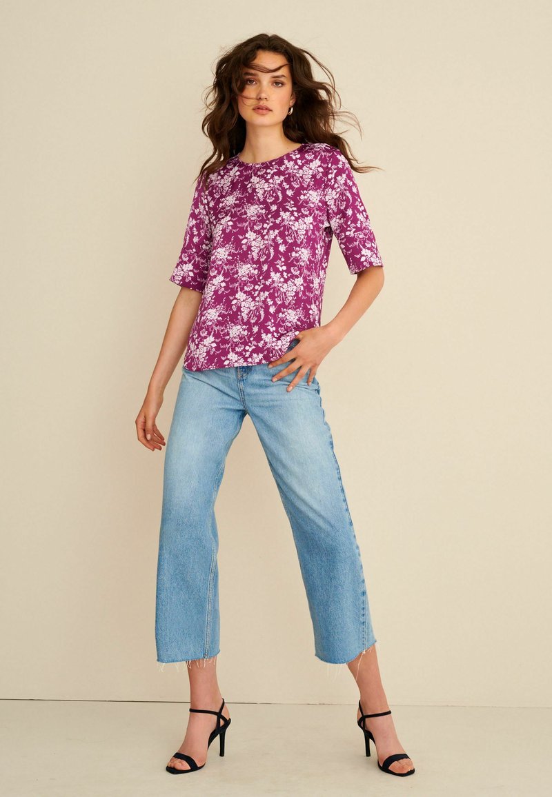Lila Oberteil mit floralem Muster und kurzen Ärmeln, kombiniert mit hellblauen weit geschnittenen Jeans und schwarzen Riemchenschuhen. Glatte Stofftextur.