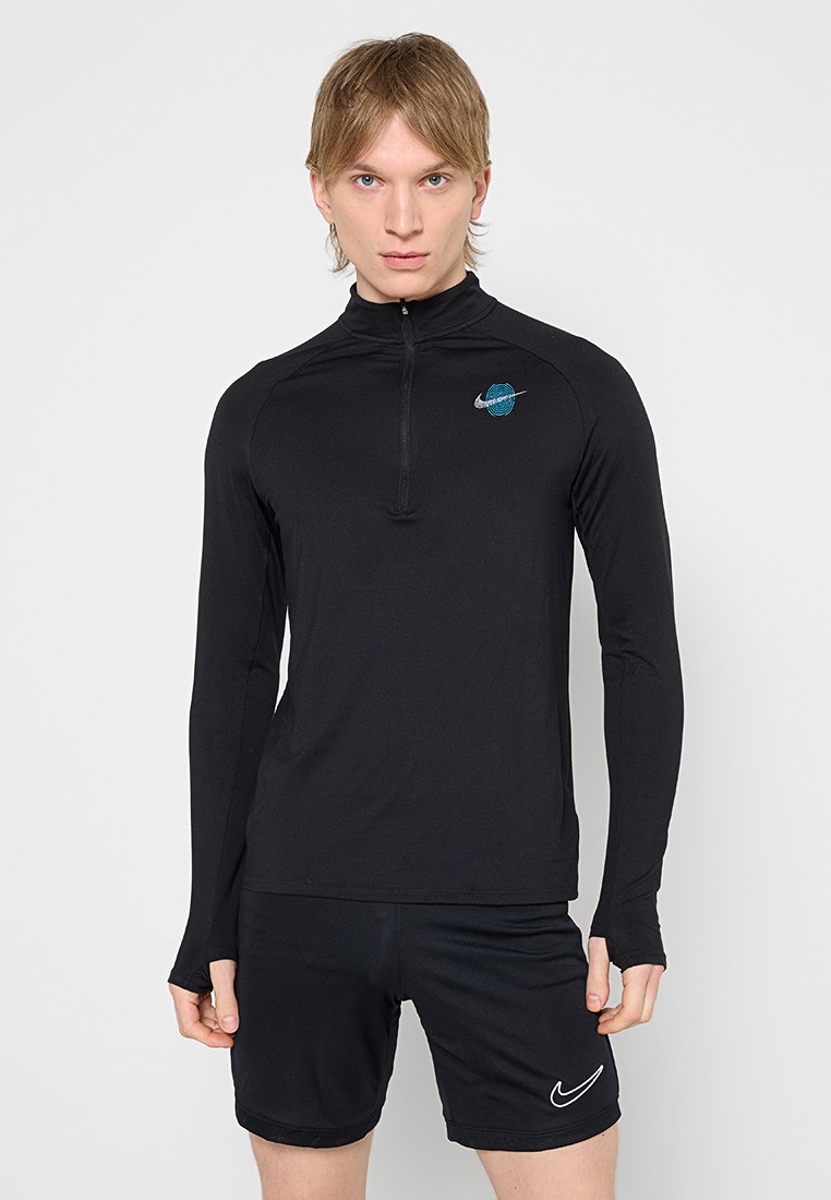 Junge Person mit blondem Haar, die ein schwarzes, langärmliges Sportoberteil mit blauem Logo und schwarze Nike-Shorts vor einem einfarbigen Hintergrund trägt.