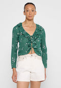 Grüne Bluse mit floralen Mustern, Rüschenkragen und Bindeband, kombiniert mit weißen Shorts. Glattes Material und taillierter Schnitt mit langen Ärmeln.