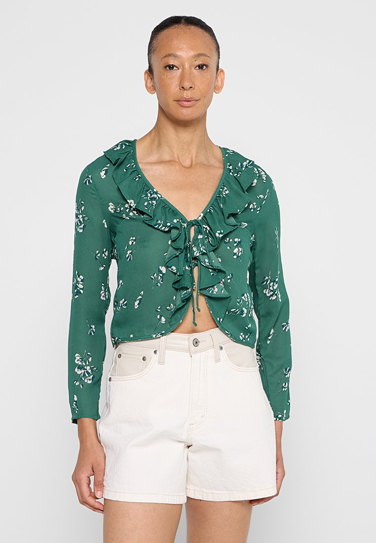 Grüne Bluse mit floralen Mustern, Rüschenkragen und Bindeband, kombiniert mit weißen Shorts. Glattes Material und taillierter Schnitt mit langen Ärmeln.