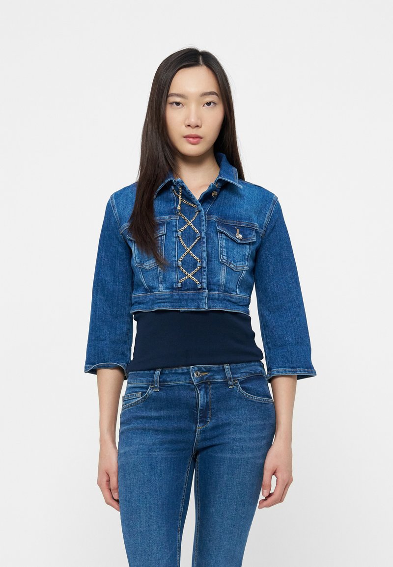 Junge Frau mit langen, glatten Haaren, die eine kurze blaue Jeansjacke mit Ketten-Schnürung und blaue Jeans trägt, stehend vor weißem Hintergrund.